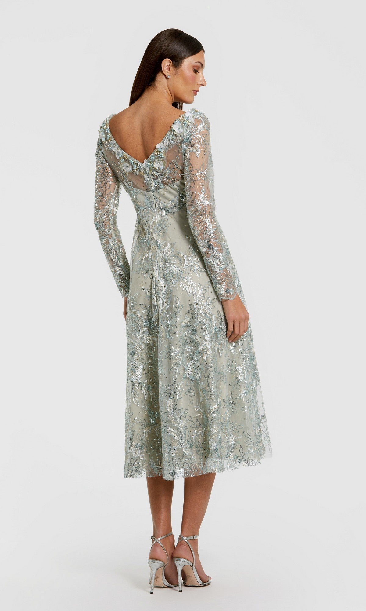 A woman stands in the Mac Duggal 68898 Midi Party Dress—a light blue, floral-embroidered midi dress with long sleeves and delicate lace details, featuring a low V-back and shimmering with subtle sequin embellishments. She completes the effortlessly elegant look with silver high heels.